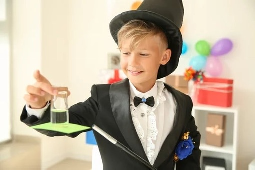 Boy dressed as a magician, balancing a glass on a card with a wand